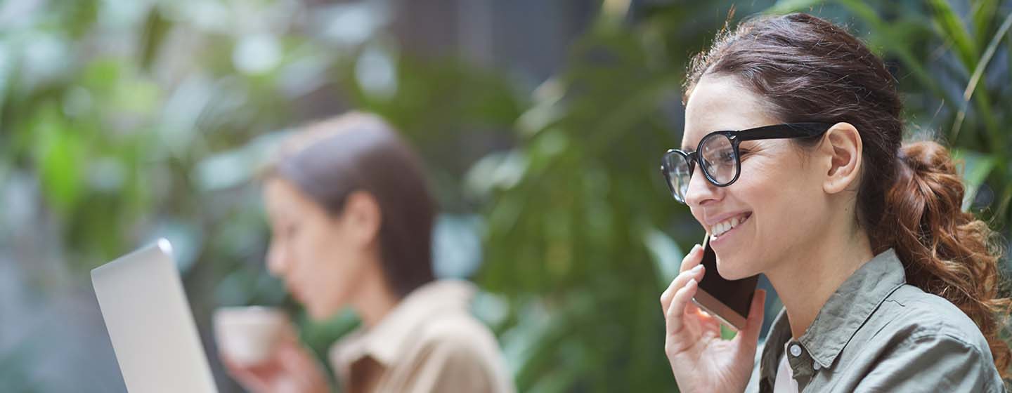 Kvinna med flexibel arbetsplats - pratar i telefon med kollega och arbetar på bärbar dator